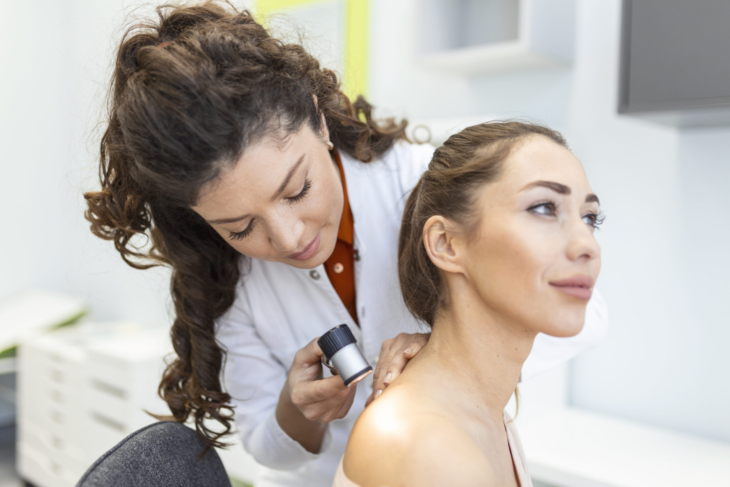 Dermatologist in latex gloves holding dermatoscope while examining attractive patient with skin disease. Female dermatologist examining patient with dermascope, looking for signs of skin cancer.